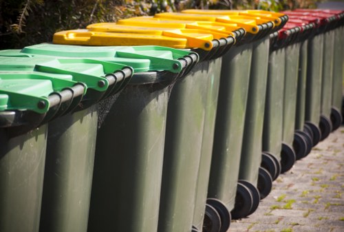 A row of large, wheeled rubbish bins lined up outdoors on a paved surface, with the nearest bin featuring a green lid, followed by bins with yellow lids, and at the end, a bin with a red lid. The bins are made of durable plastic with a smooth, slightly textured surface and are positioned on a sidewalk or driveway. In the background, there is some greenery and blurred trees, indicating an outdoor residential or community area. The image reflects typical waste collection containers used for general rubbish disposal, consistent with services provided by House Clearance Hoddesdon. The arrangement suggests readiness for waste collection, and the varying lid colours may indicate different waste types or collection purposes. The scene emphasizes the practical, organized presentation of rubbish management infrastructure common in local towns like Hoddesdon, within the postcode area, supporting efficient rubbish removal services.