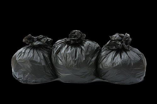 Three black garbage bags made of plastic material are positioned on a flat surface, with their tops gathered and tied. The bags appear full, with some bulges and irregular shapes indicating they contain household rubbish, possibly including items like paper, plastics, or lightweight debris. The background is dark, emphasizing the bags and their glossy surface textures, which reflect minimal lighting. The setting is likely outdoors, possibly on a driveway or pavement area near a property in Hoddesdon, as part of a rubbish removal or house clearance process carried out by House Clearance Hoddesdon. The bags are arranged in a row, with the middle bag slightly ahead, and the scene is evenly illuminated to highlight the details of the plastic material. This image exemplifies the typical visual composition of waste collection or disposal activities involving black refuse bags for household clutter clearance.