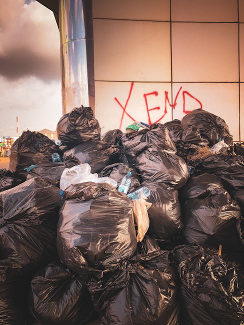 A weathered red metal skip with visible rust and scratches is situated on a dark paved ground, positioned against a modern building's grey stone and dark green panel walls. The skip's doors are slight