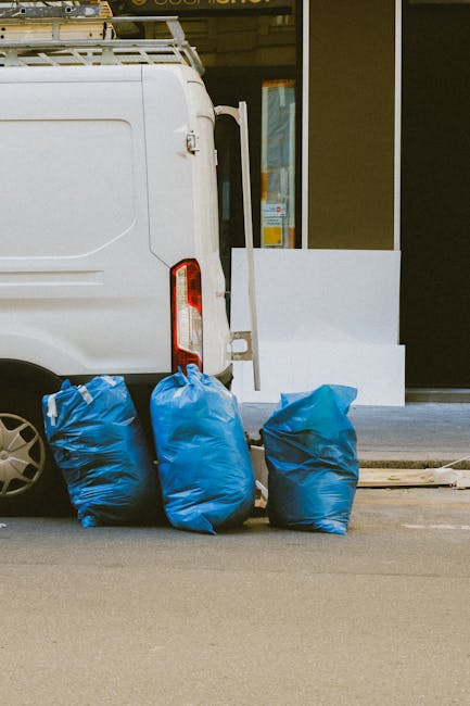 A worker wearing a dark beanie, black clothing, and a high-visibility vest stands next to a red metal trolley loaded with multiple large plastic rubbish bags in various colors, including black, grey, 